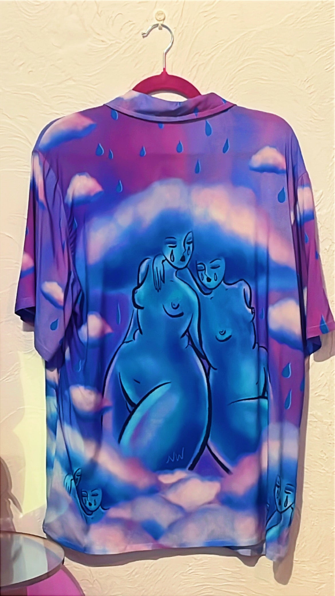 A short-sleeve button up hanging on a pink hanger against a textured beige wall. The shirt features a vibrant purple and blue  design with a central illustration of two stylized, nude figures in shades of blue and purple, surrounded by raindrop motifs. By ArtistNatalie 