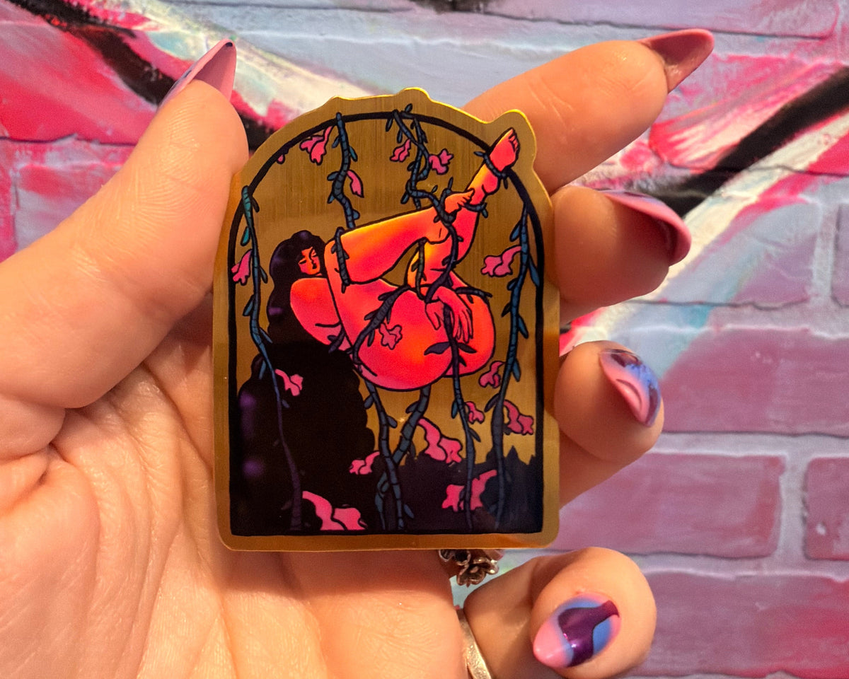 A hand with pink and blue nail art holds a small arched enamel pin featuring a stylized figure in bright pink and orange against a dark background with cherry blossom branches. The pin is set against a painted pink brick wall background.