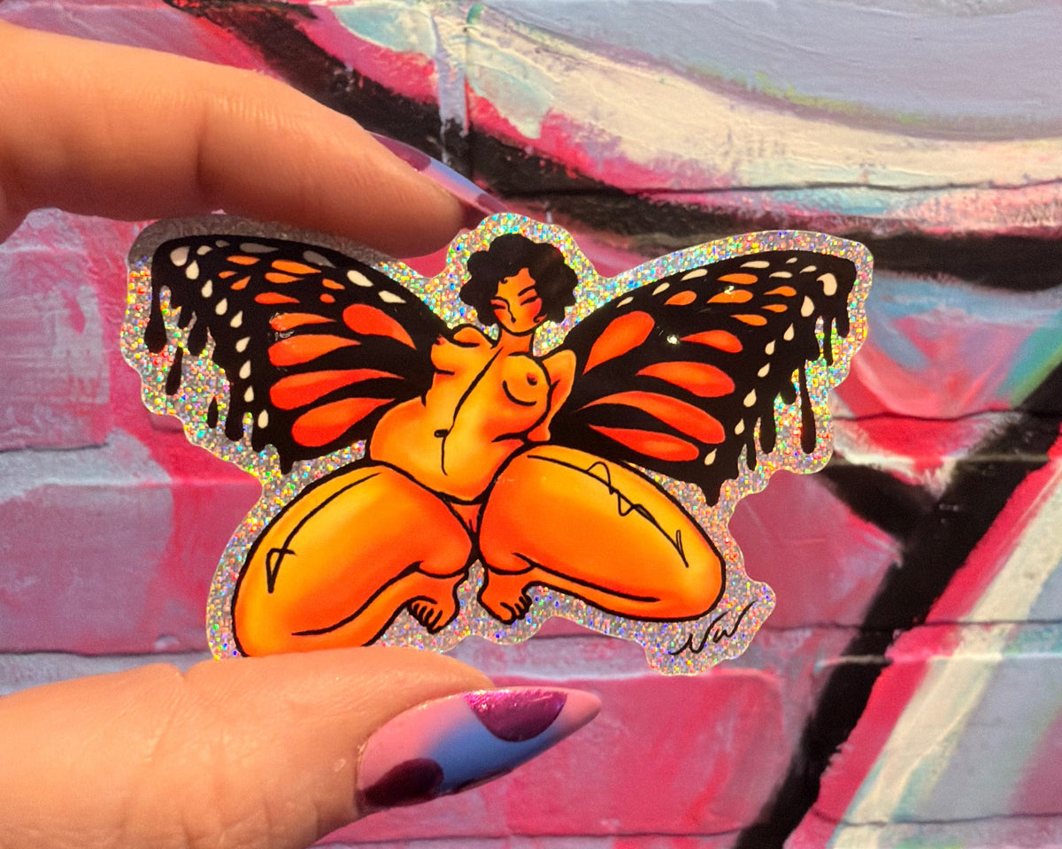 A hand with purple and blue nail polish holds a die-cut sticker featuring a stylized figure with butterfly wings in orange, black, and cream, against a colorful graffiti brick wall background.