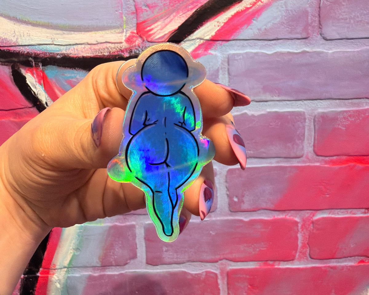 A hand with purple nail polish holds a die-cut sticker featuring a stylized pregnant figure in an astronaut suit with a holographic, iridescent blue-green finish, set against a graffiti-covered brick wall.